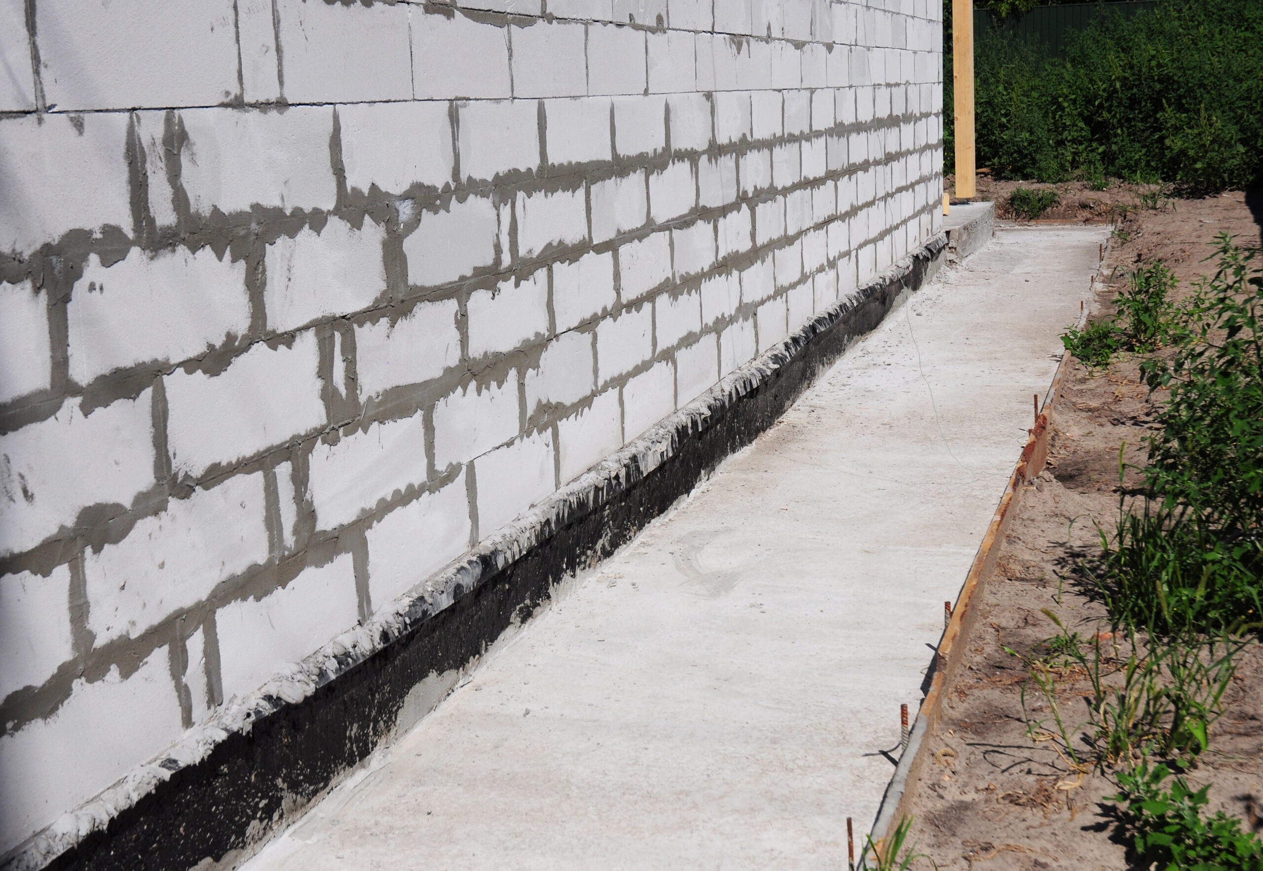 A concrete walkway runs alongside a white brick wall of a building, bordered by soil and green plants on one side.