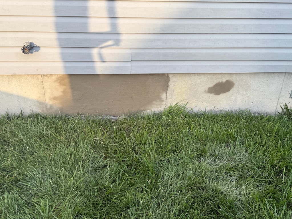 A section of foundation wall, freshly patched with cement beneath beige vinyl siding, showcases a neat cosmetic package; green grass is in the foreground.