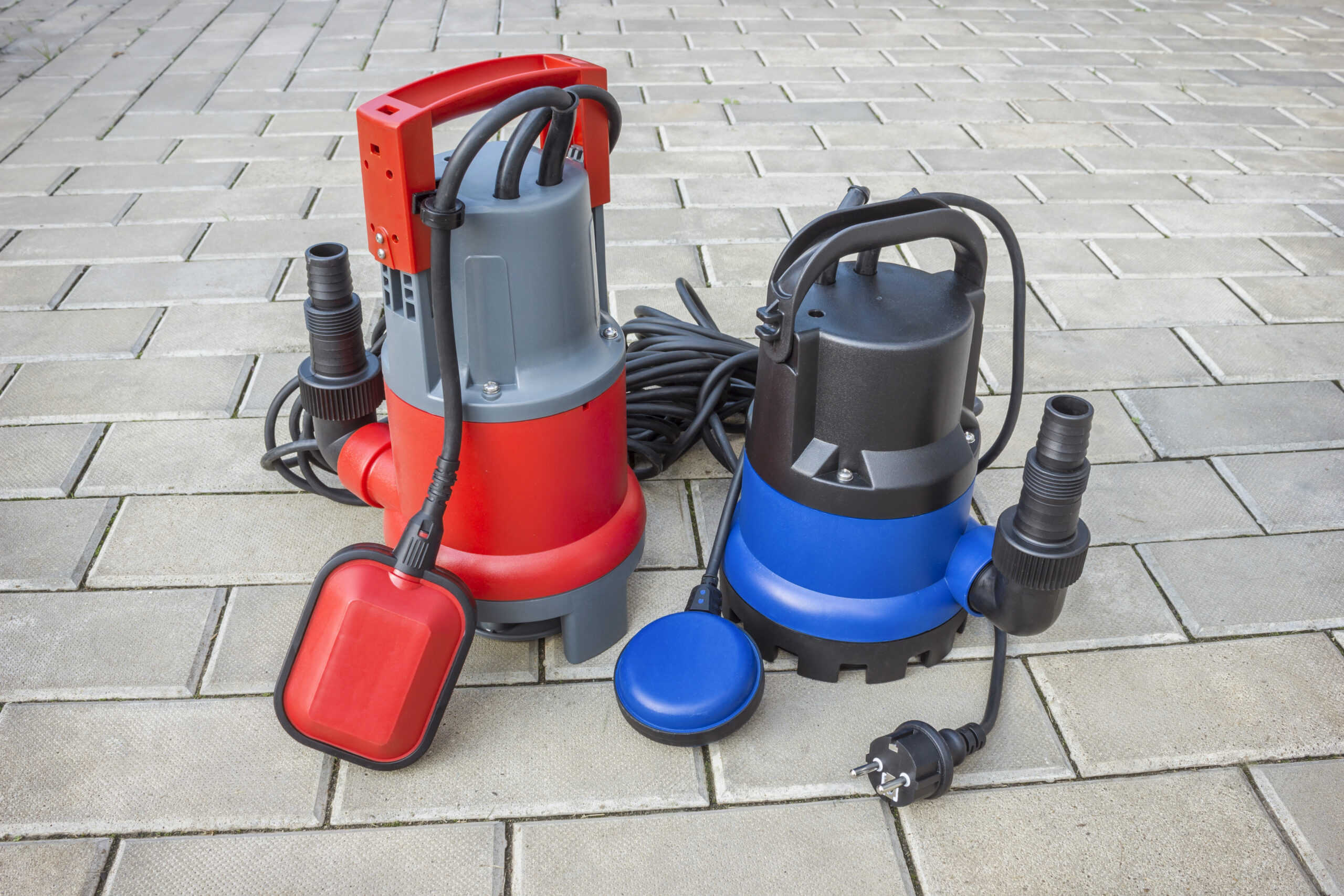 Two submersible water pumps, one red and one blue, with attached power cords and float switches, placed on a paved surface.